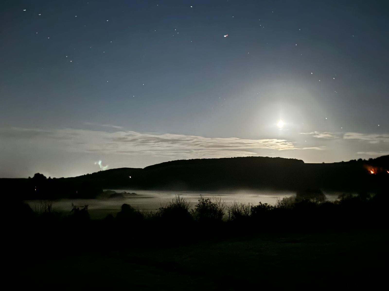 A night sky with stars and a bright moon over a landscape with hills and a body of water.