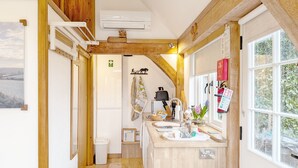 Cozinha pequena com vigas de madeira, janela com vista para o verde, pia com vários objetos e uma geladeira compacta.