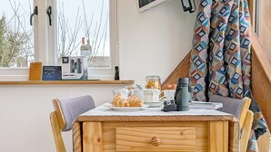 Uma mesa de jantar de madeira com toalha branca, duas cadeiras, um prato com croissants, uma cafeteira, um pote de geleia e uma câmera sobre a mesa.