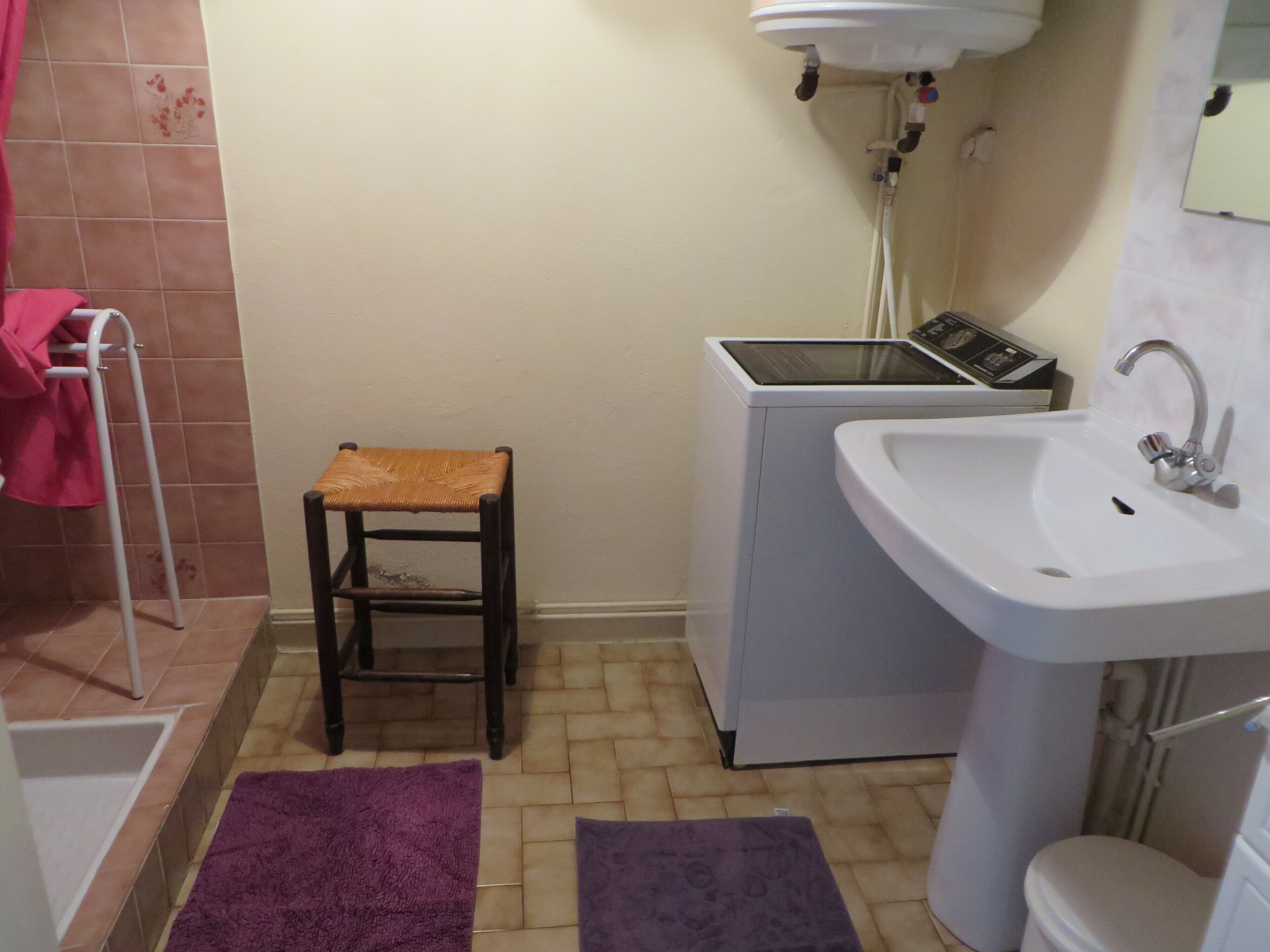 Un baño pequeño con lavadora, lavamanos, taburete y dos alfombras moradas.
