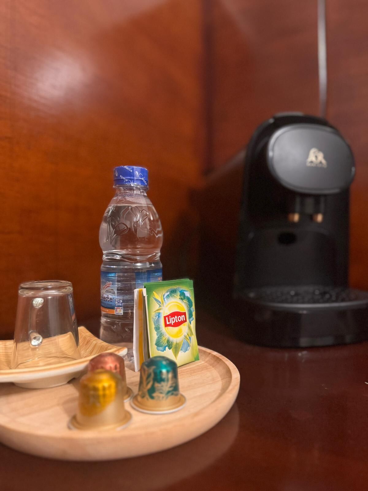 A wooden tray with a Lipton tea box, a water bottle, a coffee maker, and a glass on a table.