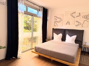A bedroom with a large bed, a wooden headboard, and a view of greenery through a sliding door. - Terre d'Eburnie Apartment Cocody Angré (Cocody)