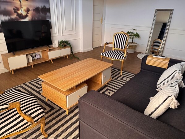 A modern living room with a wooden coffee table, striped armchair, and sofa, a flat-screen TV, and a large mirror. - Terre d'Eburnie Apartment Cocody Angré (Cocody)