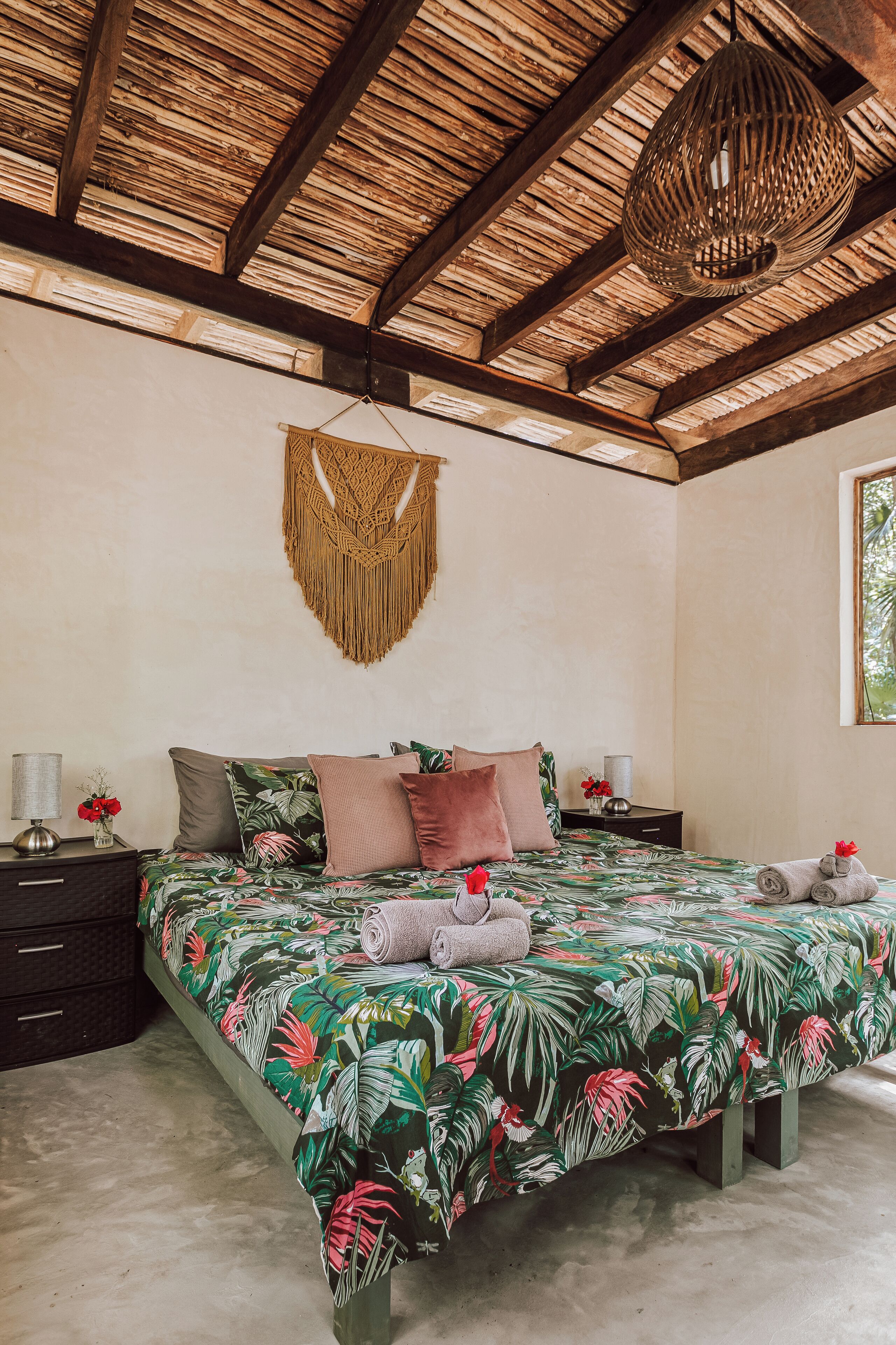 A bedroom with a bed, a hanging macrame wall hanging, a nightstand, and a window.