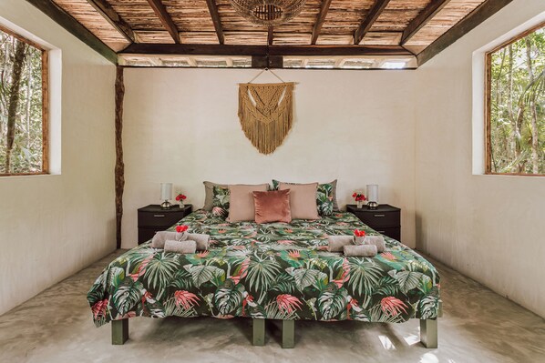 A bedroom with a bed, two bedside tables, a hanging macrame wall decoration, and large windows with a view of trees. - Casa Arkaana Eco Retreat (Tulum)