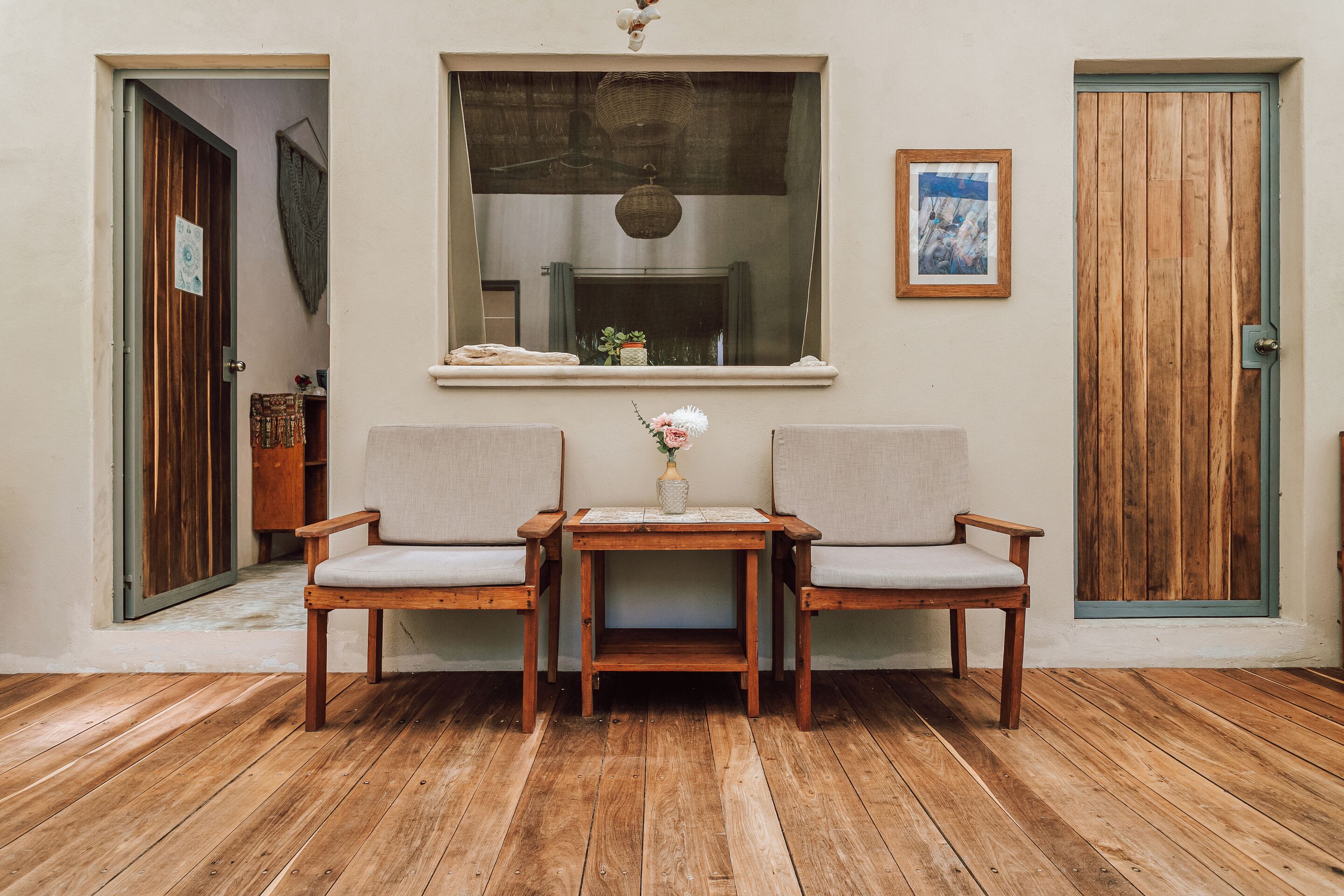 A room with wooden flooring, two chairs, a small table with a vase, a window with a view, and a door.