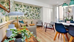 A modern living room with a wooden floor, a dining area with a white table and blue chairs, and a wall with tropical leaf wallpaper. - Raffinato appartamento con piccolo giardino al Vaticano  (ROMA)