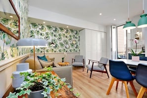 Ein modernes Wohnzimmer mit Holzboden, einem Essbereich mit weißem Tisch und blauen Stühlen sowie einer Wand mit tropischem Blattmuster-Tapete.