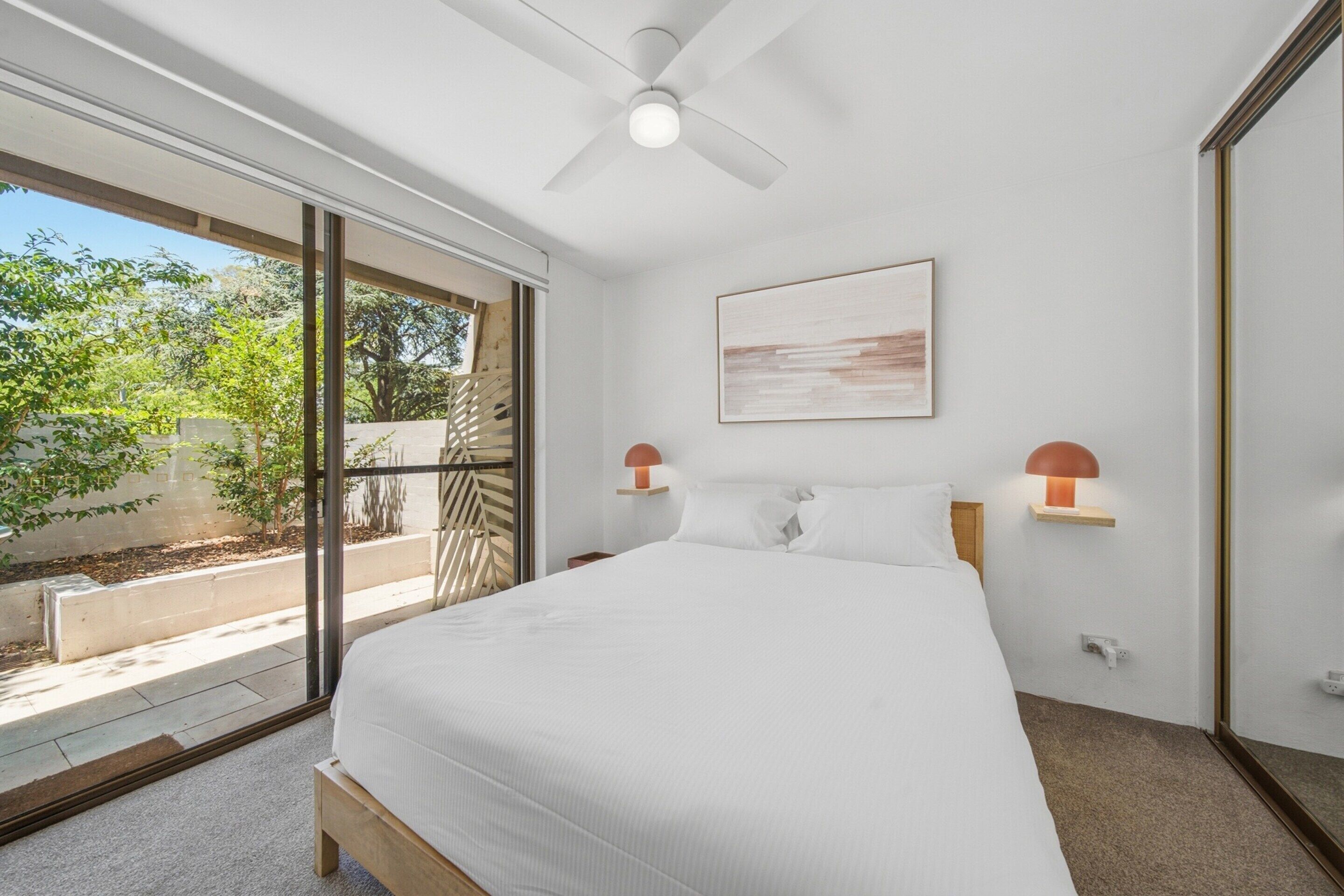 A bedroom with a large bed, a ceiling fan, a window with a view of greenery, and two bedside lamps.