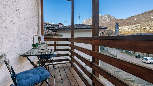Un balcon doté d’une balustrade en bois, une chaise bleue avec un coussin, une petite table sur laquelle est posée une plante en pot, et une vue sur une ville avec un clocher d’église et des montagnes.