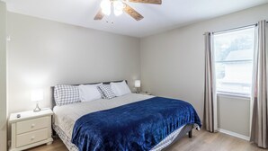 Ein Schlafzimmer mit Bett, Nachttisch, Lampe und einem Fenster mit Vorhängen.