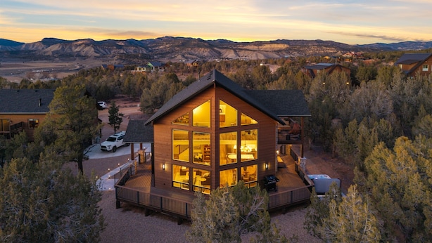A modern log cabin with a large glass facade, a deck, and a hot tub, surrounded by a forest and mountains in the background. - Zion East Luxury | Private Hot Tub & Fire Pit (Orderville)