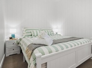 Un lit à baldaquin blanc avec une literie à rayures vertes et blanches, une table de chevet blanche avec une lampe et un plaid en tissu tissé.