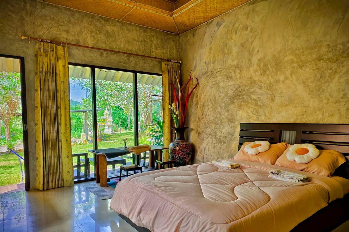 Une chambre à coucher avec un grand lit, une tête de lit en bois et une vue sur un jardin grâce à de grandes fenêtres.