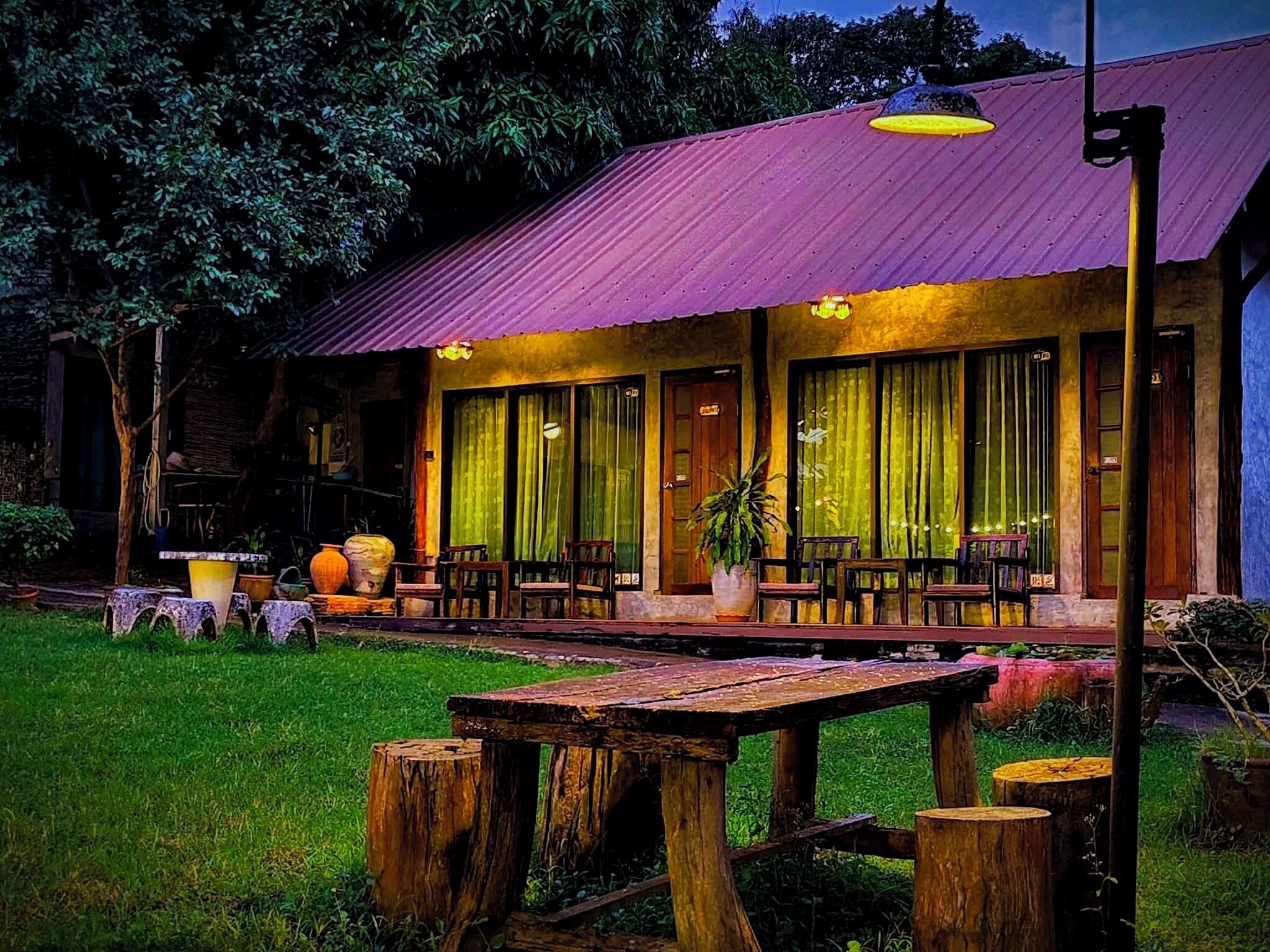 A rustic building with a metal roof and large windows, surrounded by a grassy area with wooden tables and chairs.