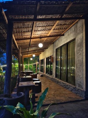 An outdoor patio with stone flooring, wooden tables, and chairs, surrounded by large windows and a pergola with string lights. - Phakphing Erawan Homestay (Tha Kradan)
