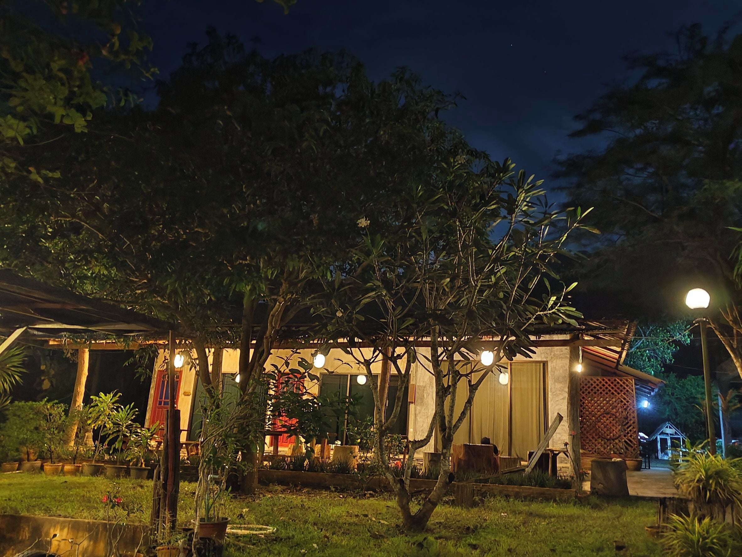 Un espacio al aire libre iluminado por la noche con un área de estar cubierta, plantas en macetas y una fuente de agua pequeña.