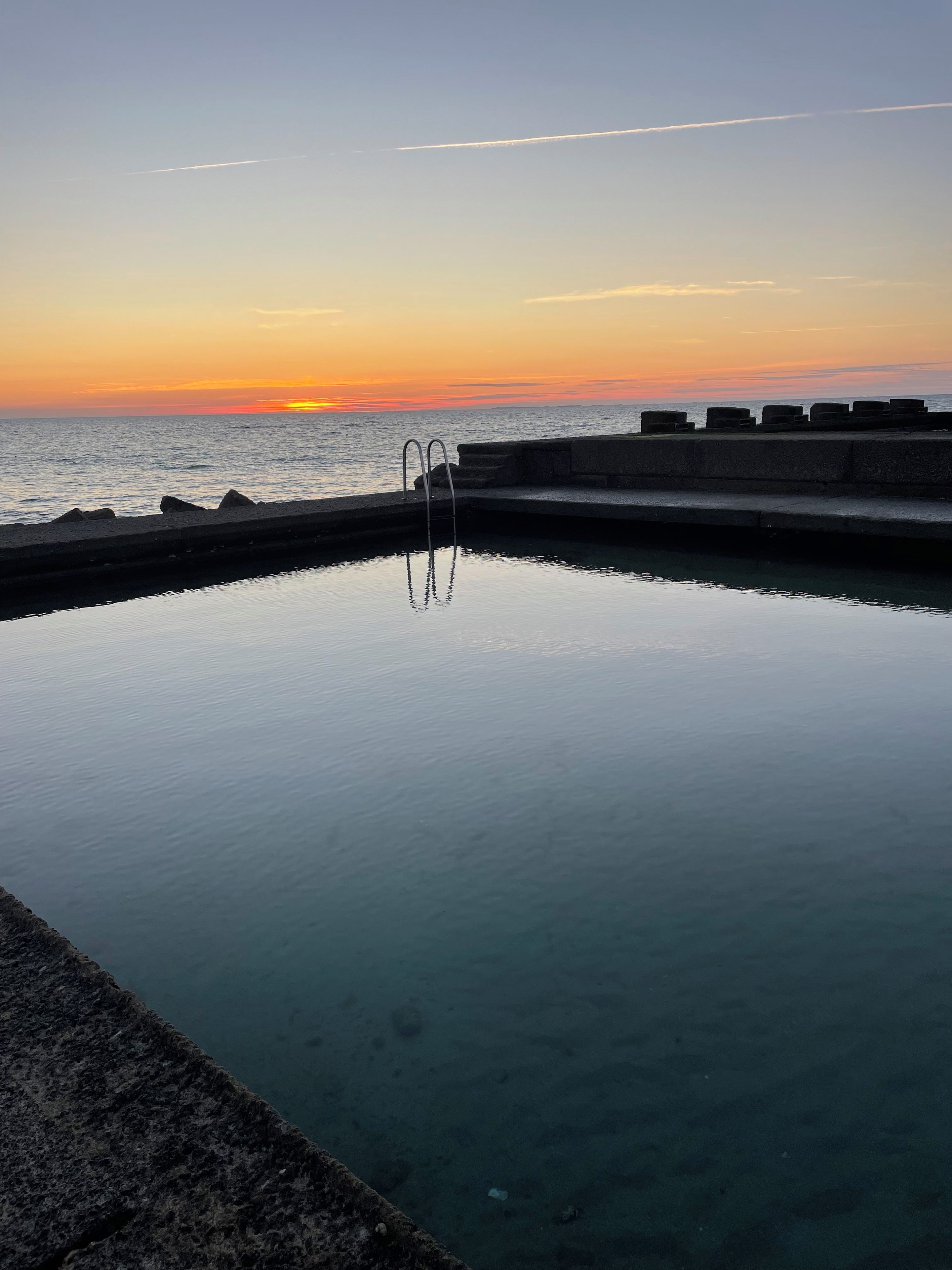 A swimming pool with a concrete edge, a metal ladder, and a view of the ocean at sunset.