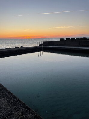 A swimming pool with a concrete edge, a metal ladder, and a view of the ocean at sunset. - Apartment with a view of the ocean (Donville-les-Bains)
