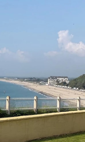 A coastal view with a beach, a fence, and a building in the distance. - Apartment with a view of the ocean (Donville-les-Bains)