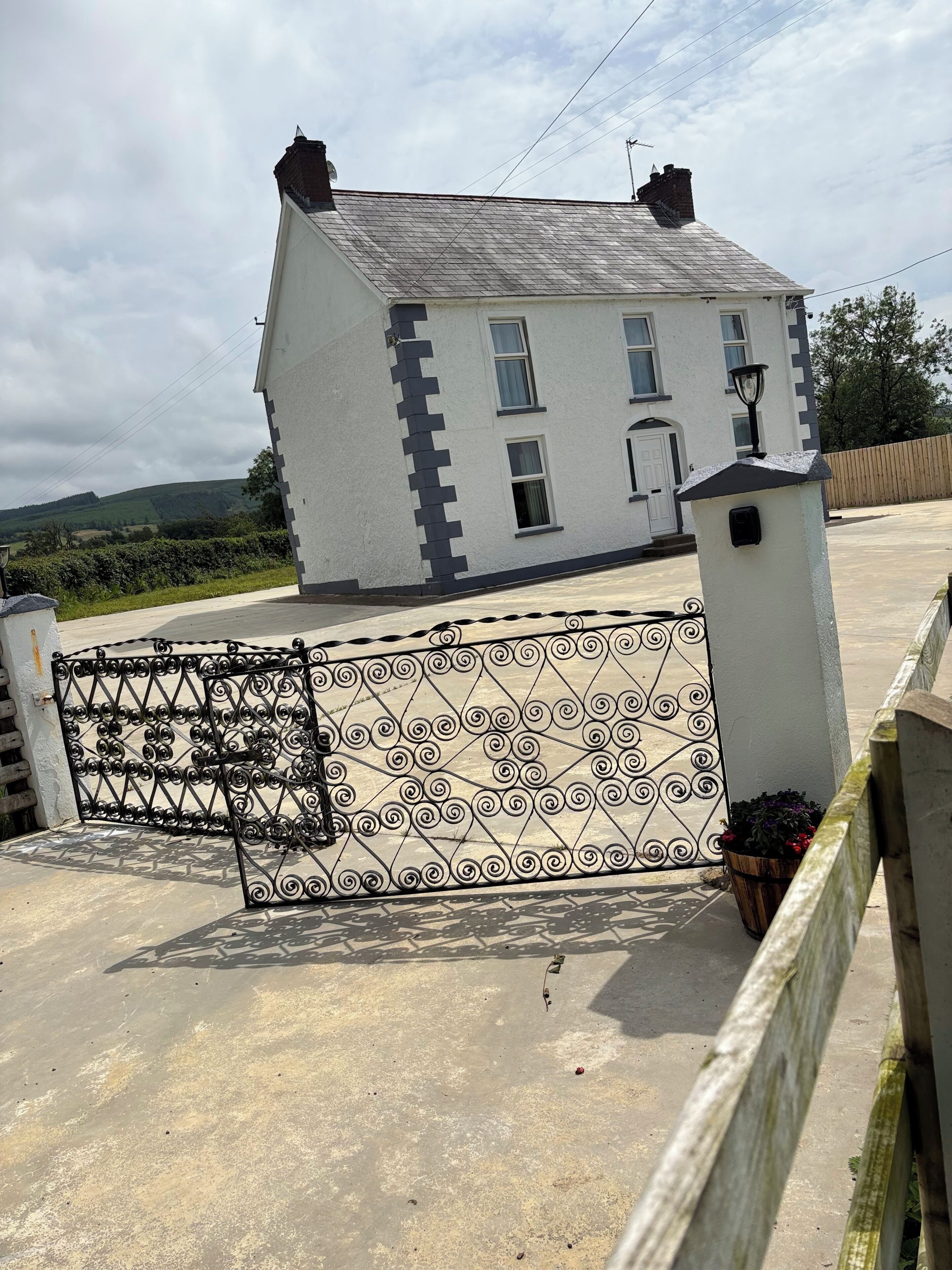 Une maison à deux étages, avec une façade blanche et un toit gris, une entrée fermée par un portail et un petit jardin agrémenté de plantes en pot.