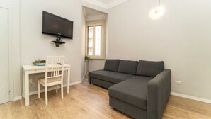 Een kamer met een grijze bank, een wit bureau met een stoel, een wandtelevisie en een raam met uitzicht op gebouwen.