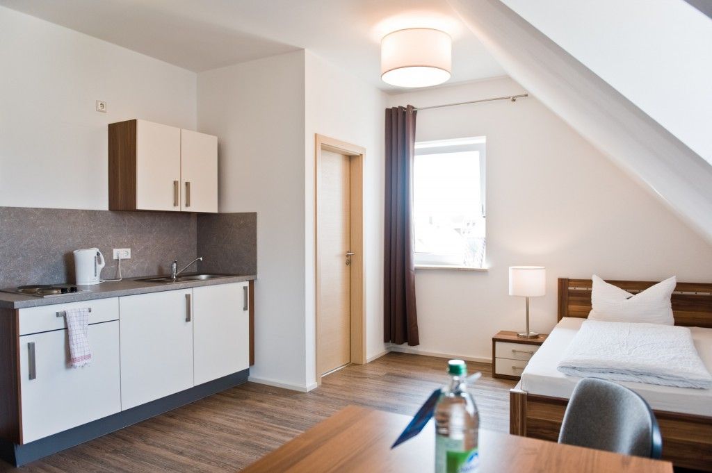 Habitación de hotel pequeña con kitchenette, una cama, una mesita de noche y un ventanal con cortinas.