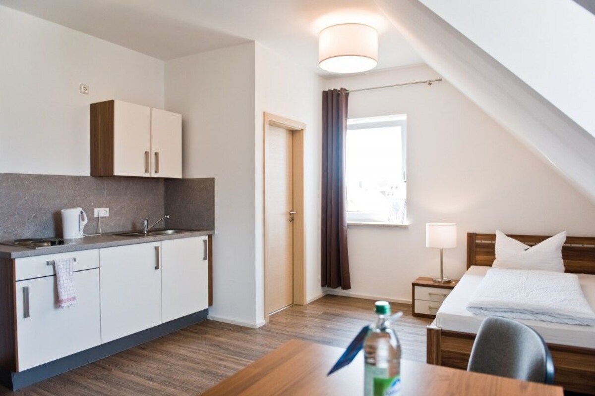 Habitación de hotel pequeña con kitchenette, una cama, una mesita de noche y un ventanal con cortinas.
