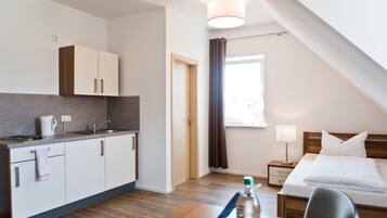 Habitación de hotel pequeña con kitchenette, una cama, una mesita de noche y un ventanal con cortinas.