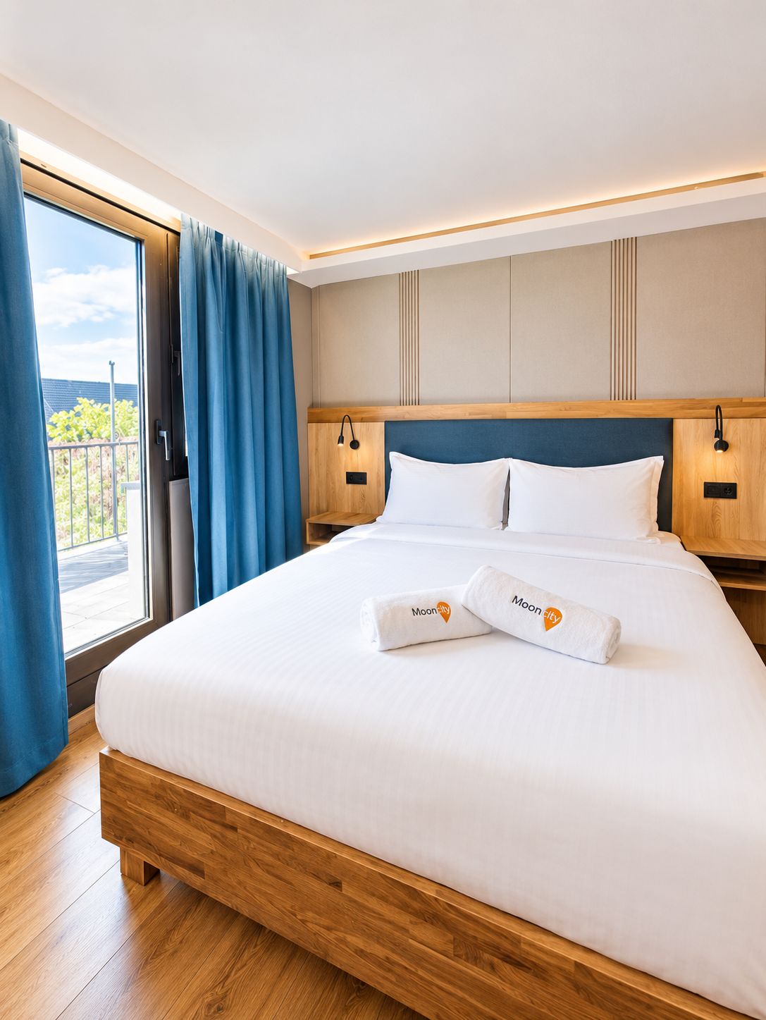 Une chambre d’hôtel avec un grand lit, une tête de lit en bois et une vue sur un balcon extérieur accessible par une porte coulissante.
