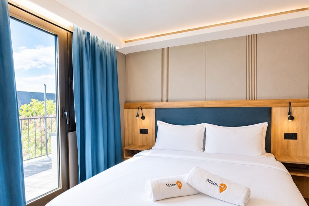 A hotel room with a large bed, wooden headboard, and a view through a sliding door to an outdoor balcony.