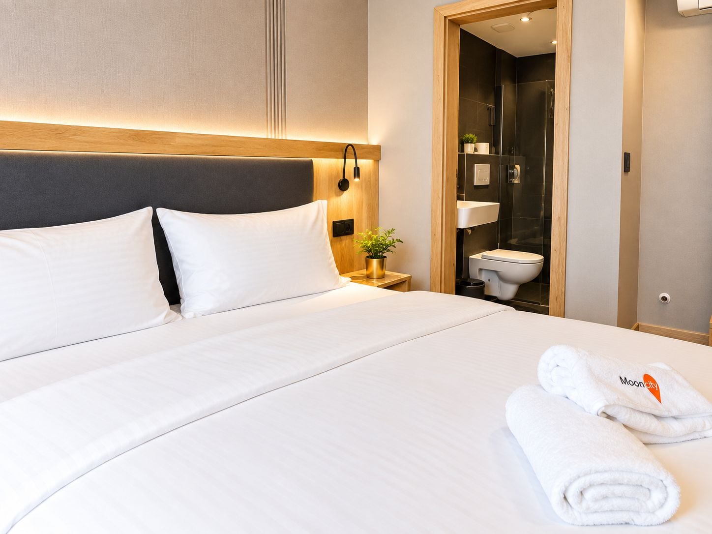 Una habitación de hotel con cama, mesita de noche, planta en maceta y baño visible a través de una puerta abierta.
