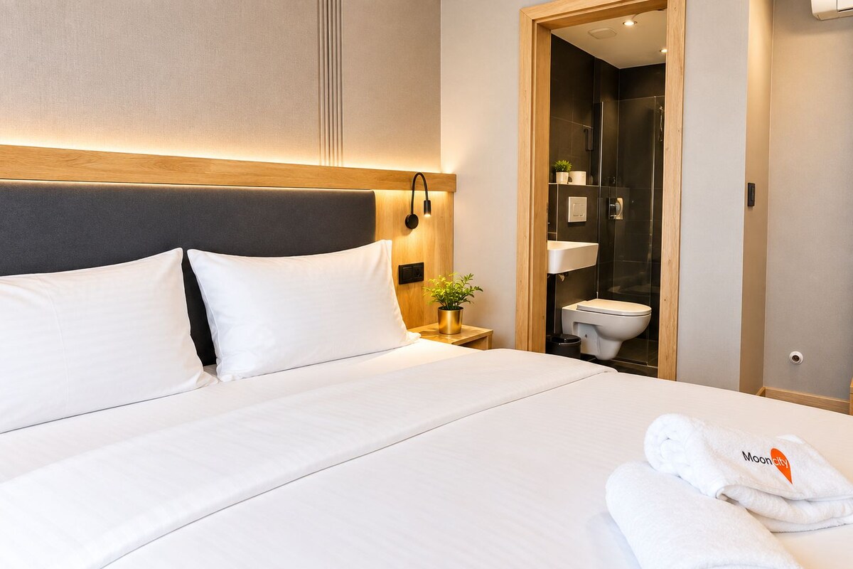 Una habitación de hotel con cama, mesita de noche, planta en maceta y baño visible a través de una puerta abierta.