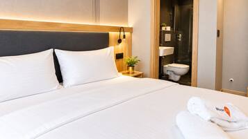 Una habitación de hotel con cama, mesita de noche, planta en maceta y baño visible a través de una puerta abierta.