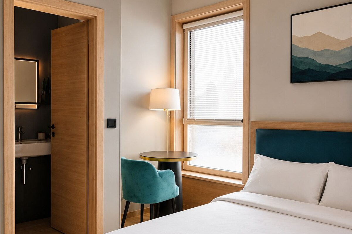 Habitación de hotel con cama, escritorio, silla, lámpara, ventana con persianas y un cuadro en la pared.
