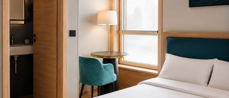 A hotel room with a bed, a desk, a chair, a lamp, a window with blinds, and a painting on the wall.