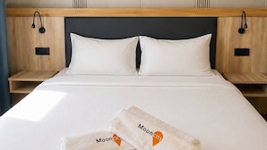 A neatly made bed with white linens and two towels with 'MoonCity' branding, a wooden headboard, and a wall with vertical lines. - Mooncity Hotel & Boardinghouse  (Düsseldorf)