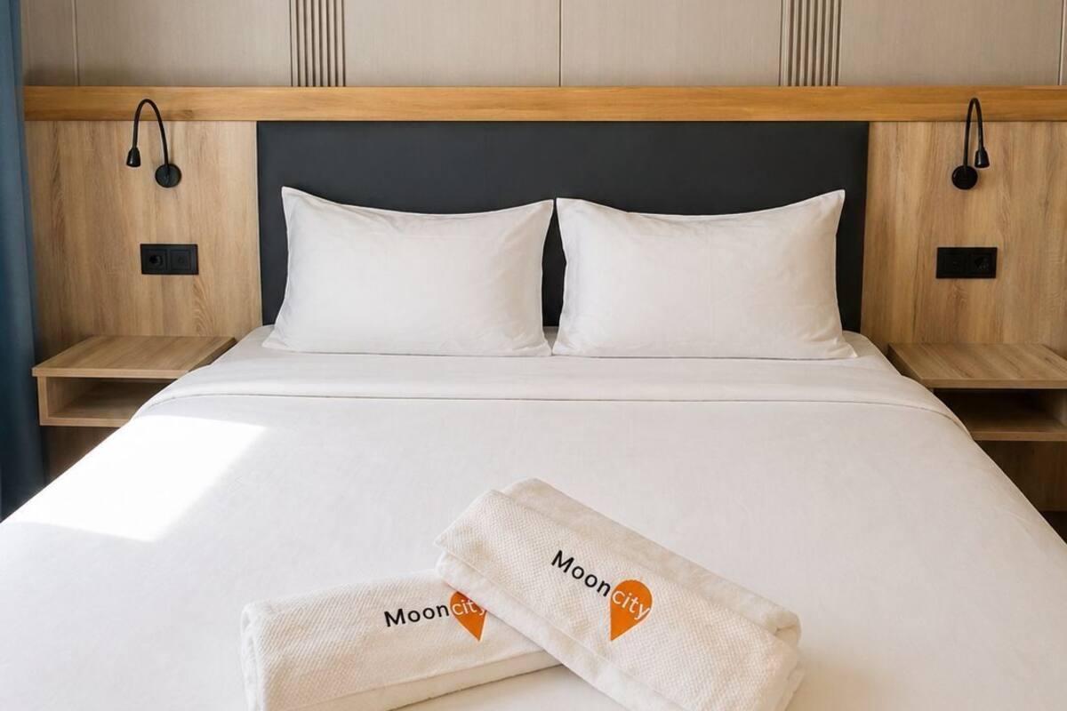 Un lit bien fait, avec du linge de lit blanc et deux serviettes ornées du logo « MoonCity », une tête de lit en bois et un mur décoré de lignes verticales.