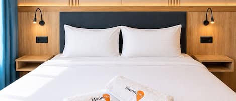 Habitación de hotel con una cama grande, dos almohadas, dos mesitas de noche con lámparas y un par de toallas con un logo.