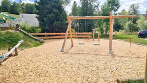 A playground with swings and a slide, surrounded by a wooden fence and trees. - "Fewo Fox'n" – The Clever Accommodation with Pool (Schluchsee)