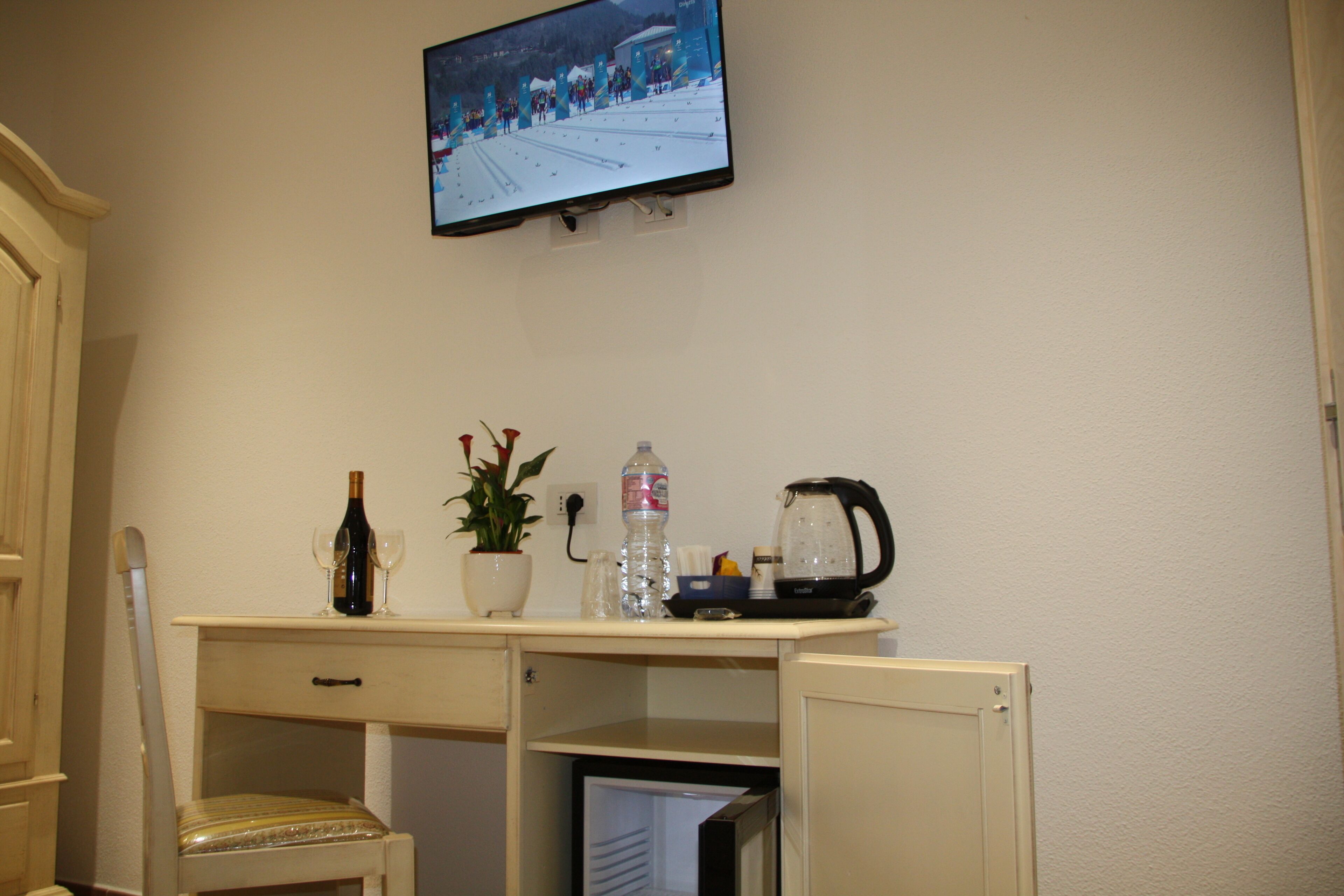 A hotel room with a desk, a chair, a television mounted on the wall, a bottle of wine, a water bottle, a plant, and a coffee maker.