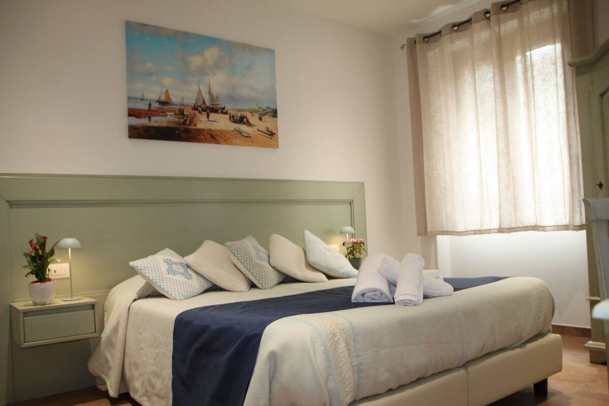 A neatly made bed with a blue and white bedspread, a painting of a beach scene above it, and a window with sheer curtains.