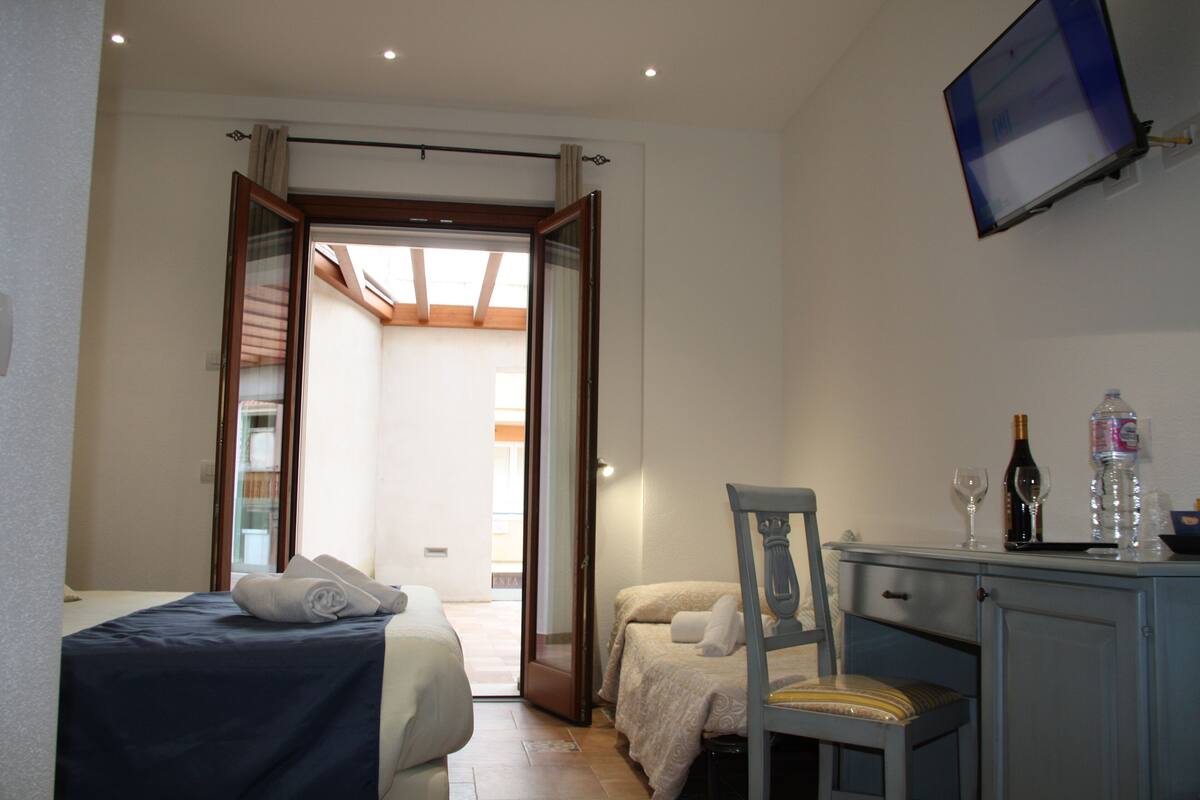 A hotel room with a bed, a desk with a chair, a television mounted on the wall, and a door leading to an outdoor area.