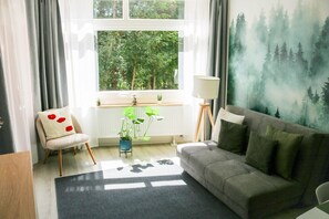 Un salon comprenant un canapé, un fauteuil, un radiateur, une fenêtre donnant sur des arbres et une lampe.