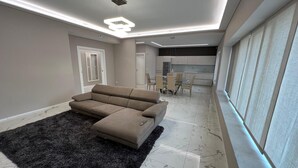 A modern living room with a beige sectional sofa, a black rug, and a kitchen in the background. - Turin Urban Marble Suite (TORINO)