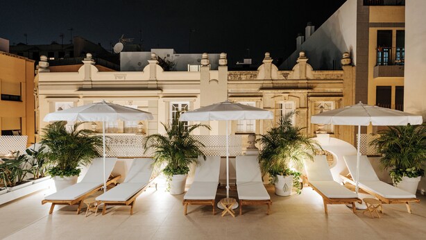 Un área de lounge en la azotea con tumbonas blancas y sombrillas, plantas en macetas y un edificio con arquitectura clásica al fondo.