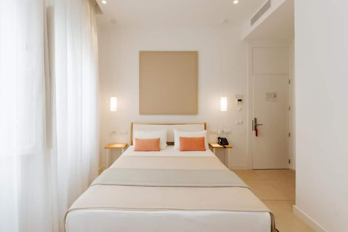 Une chambre d’hôtel avec un grand lit, deux tables de chevet, une petite table, une lampe fixée au mur et une porte.