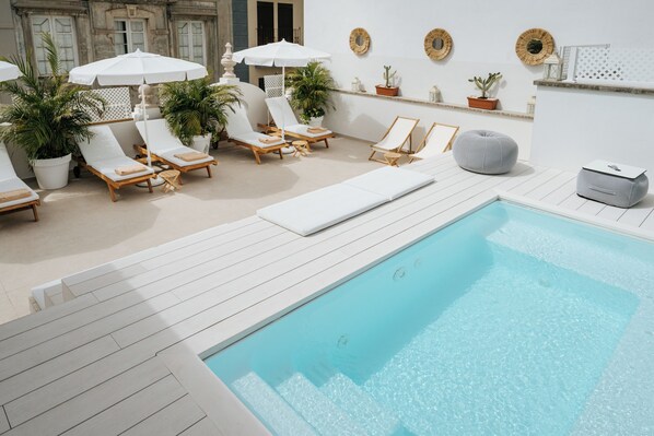 Zona junto a la piscina con sillones, sombrillas, plantas en macetas y un pequeño rincón de descanso.