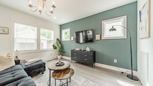 A living room with a grey sofa, a coffee table, a TV, and a plant. - Spacious Home-6 Beds | 10 Mins From City Center (Charlotte)