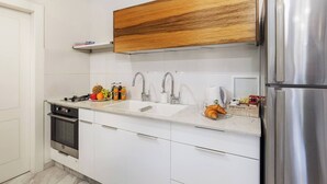 A modern kitchen with white cabinets, a stainless steel refrigerator, a wooden upper shelf, and a sink with a faucet. - 1 BR Apartment in Schneller Jerusalem Israel (Jerusalem)